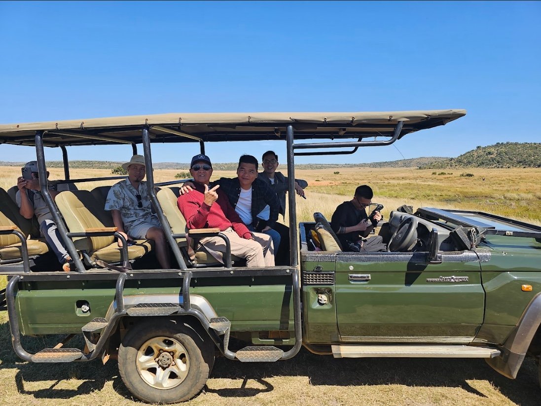 Nhóm người chụp ảnh trong xe safari tại một khu bảo tồn thiên nhiên, với phong cảnh đồng cỏ rộng lớn phía sau.