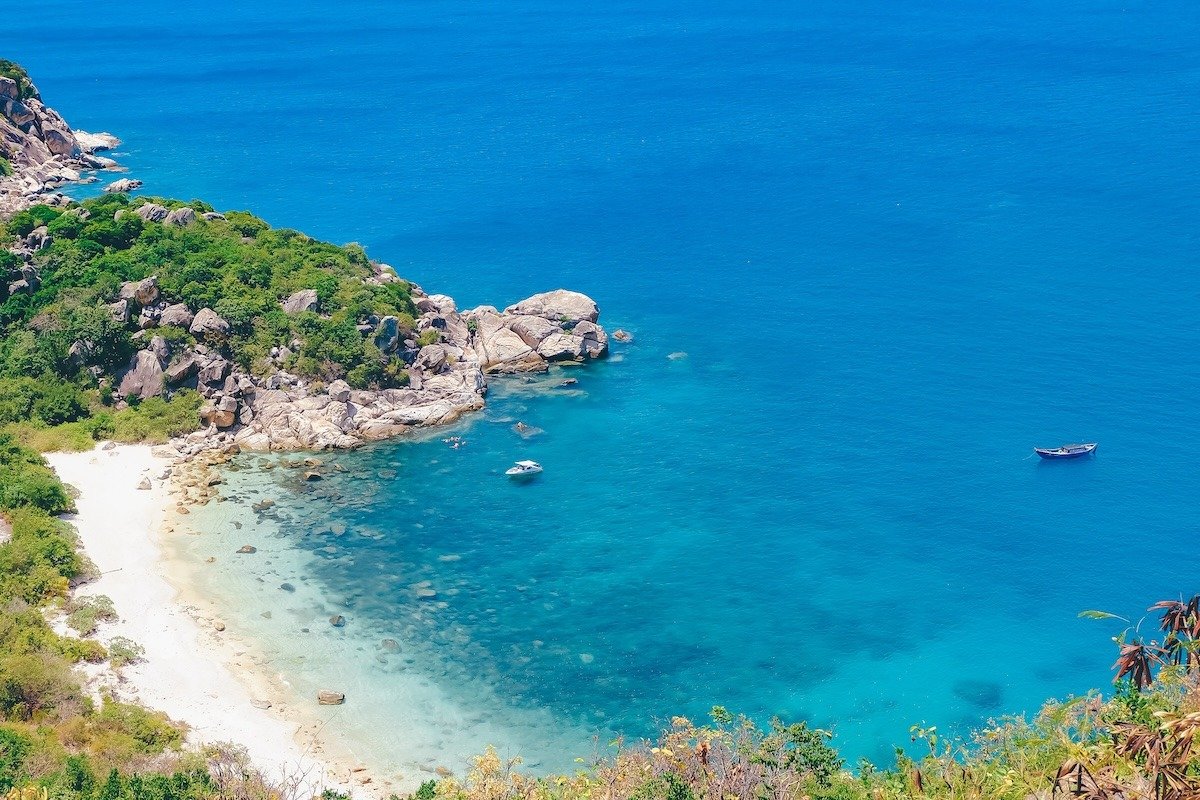 Bãi biển tuyệt đẹp với cát trắng, nước biển trong suốt, thuyền nhỏ và bờ đá phủ cây xanh tươi.