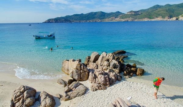 Một bãi biển với nước biển xanh trong, bãi đá, và du khách đang bơi lội, tàu thuyền neo đậu xa xa.