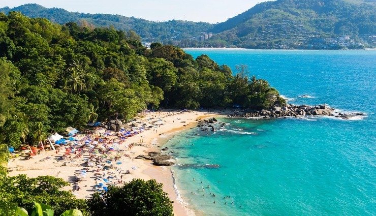 Bãi biển nhiệt đới xanh trong với du khách thư giãn dưới ô che nắng, núi đồi xanh tươi vây quanh.