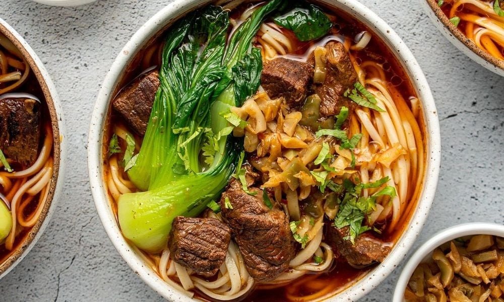 Bát mì thịt bò cay nóng hổi, kết hợp cải thìa tươi xanh, đặt trên nền bàn xám, hấp dẫn khẩu vị.