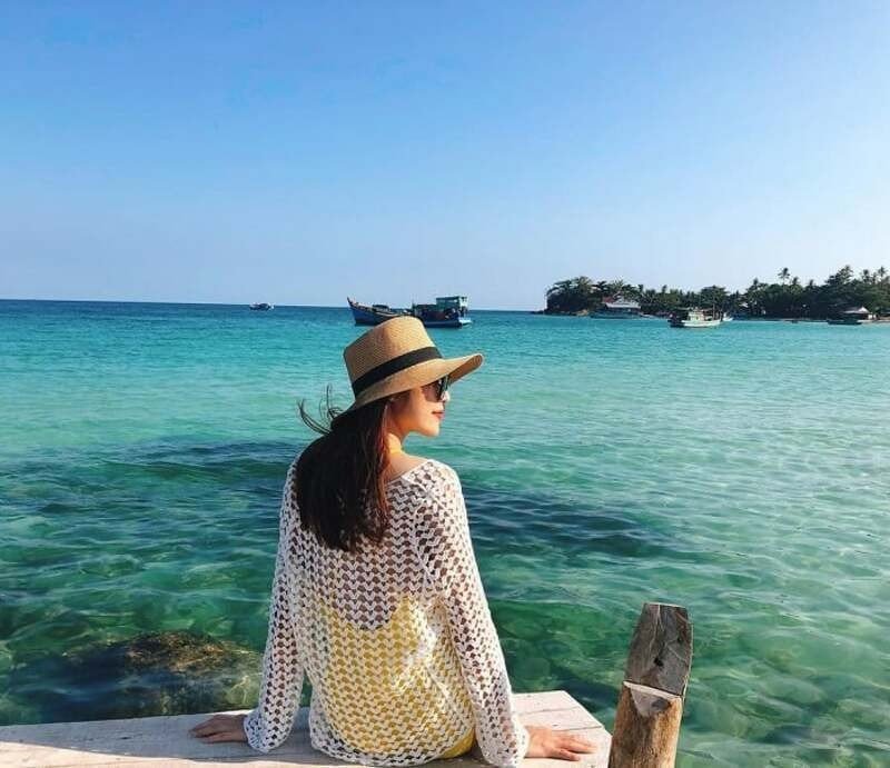 Người phụ nữ ngồi trên bến tàu, đeo mũ rộng vành, ngắm biển xanh và những hòn đảo xa xôi vào ngày nắng đẹp.