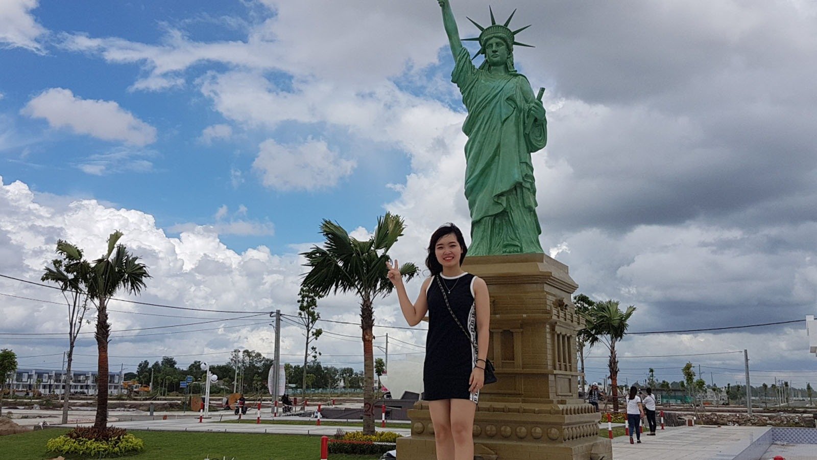 Người phụ nữ đứng bên bức tượng Nữ thần Tự do mini trong khuôn viên có trời mây xanh.