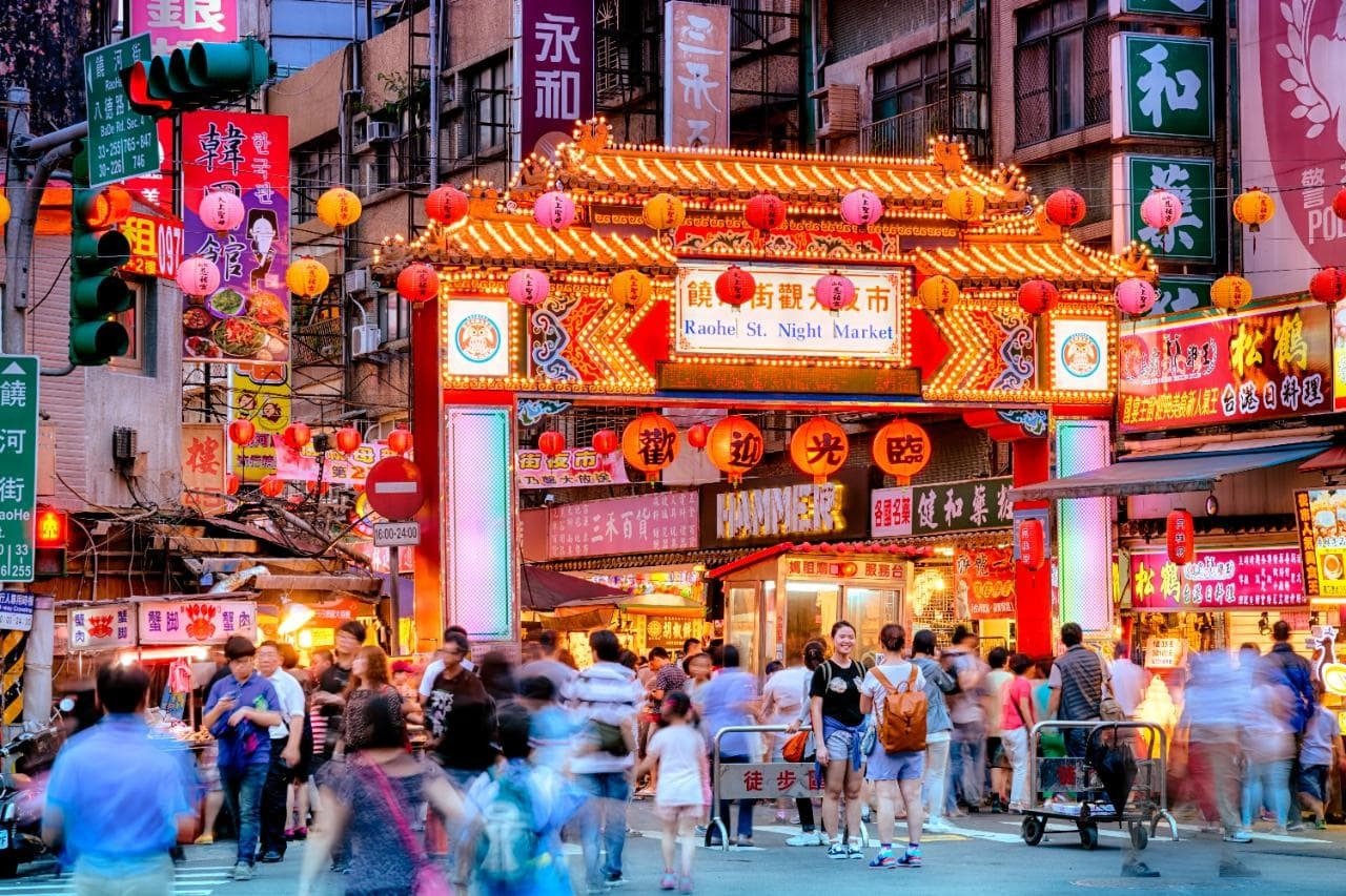 Đám đông đi bộ tại cổng vào chợ đêm sôi động với đèn lồng và biển hiệu sáng rực, khu phố nhộn nhịp buổi tối.