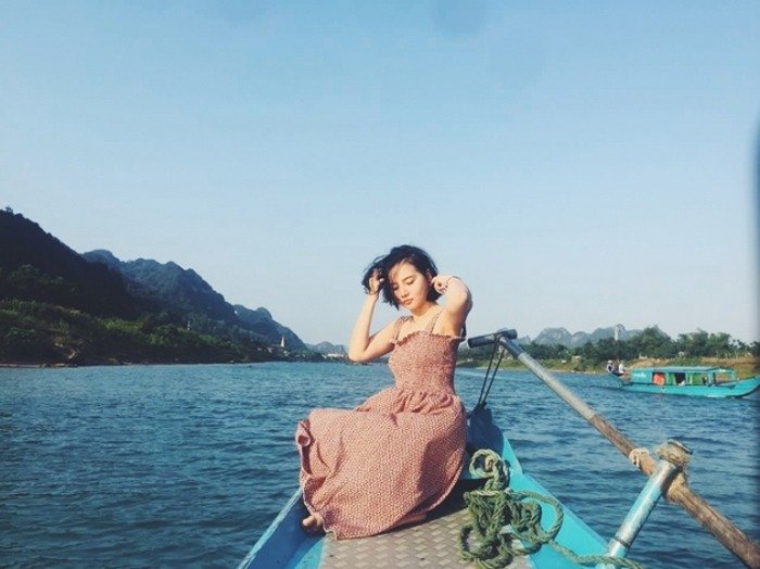 Người phụ nữ ngồi thư giãn trên mũi thuyền giữa dòng sông xanh, trời trong xanh và núi đồi phía xa.