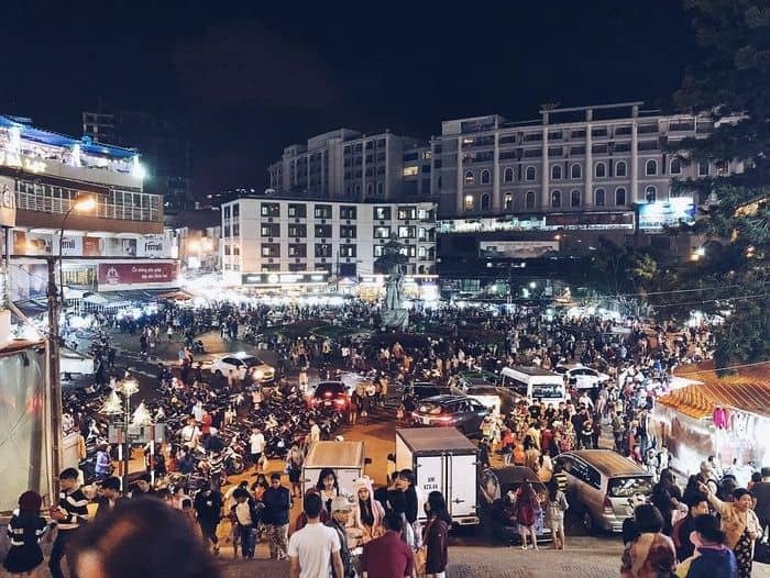 Chợ đêm Đà Lạt sôi động với đám đông người tham quan, các quầy hàng sáng đèn và tòa nhà xung quanh, tạo khung cảnh nhộn nhịp.