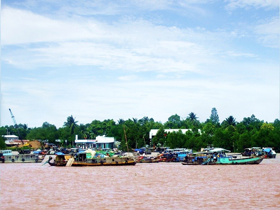 Thuyền đánh cá neo đậu trên dòng sông, bên bờ là dãy nhà cửa và cây cối xanh tươi dưới bầu trời xanh.