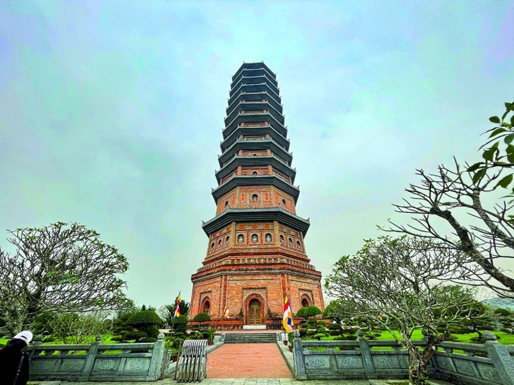 Tháp tôn giáo bằng gạch cao nổi bật giữa sân vườn xanh tươi, bầu trời xám nhạt phía trên, cây cối xung quanh.