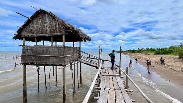 Căn chòi gỗ mộc mạc bên bờ biển, đường đi bằng gỗ, trời nắng, người dân đi dạo trên bãi cát.