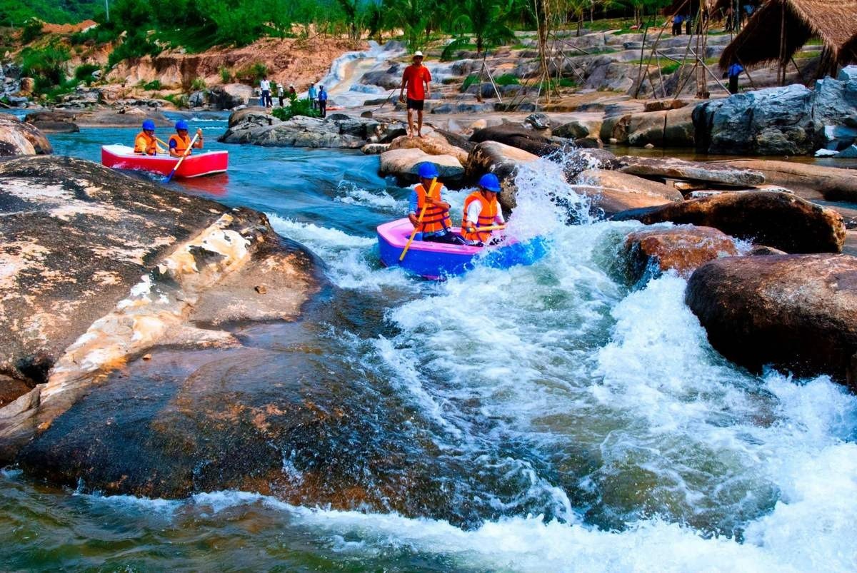Trẻ em chèo thuyền kayak xuống dòng suối với áo phao và mũ bảo hiểm, xung quanh là cảnh quan thiên nhiên hoang sơ.