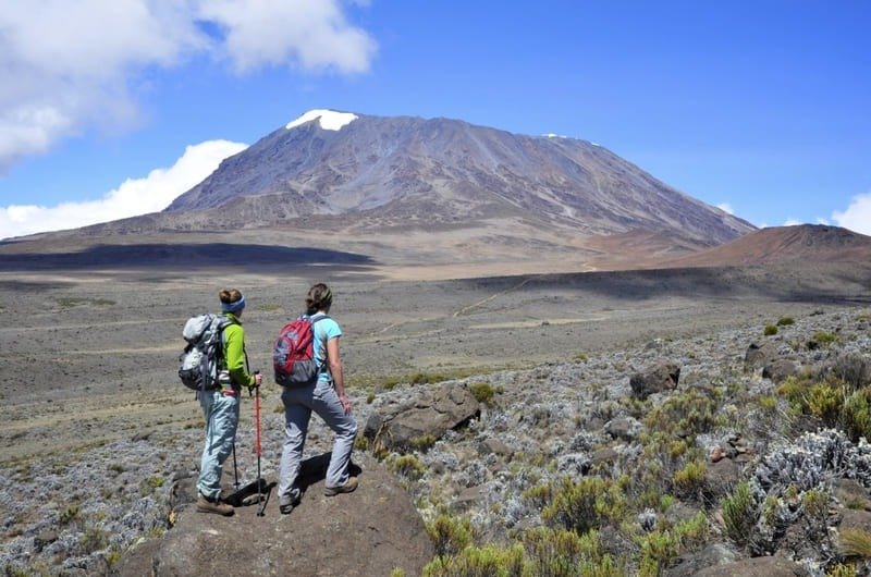 Hai người leo núi đang ngắm cảnh núi Kilimanjaro hùng vĩ dưới bầu trời xanh mây trắng.