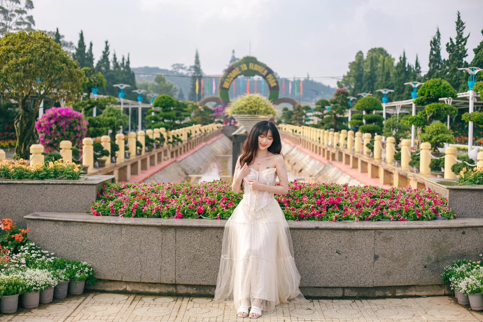 Người phụ nữ đứng trước vườn hoa rực rỡ trong công viên, mặc váy trắng, mỉm cười đầy thư thái và hạnh phúc.
