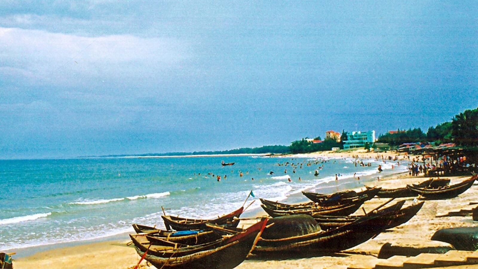 Bãi biển với tàu thuyền gỗ và du khách vui chơi dưới ánh nắng, đường chân trời xanh thẳm xa xa.