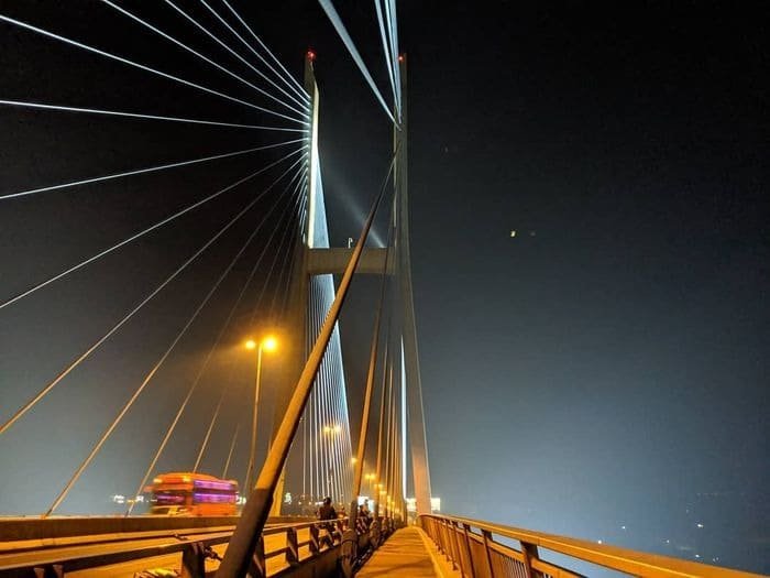 Cầu dây văng lung linh trong ánh đèn đêm, với góc nhìn từ làn đường bộ và xe cộ đang di chuyển.