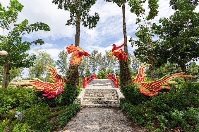 Cổng chào trang trí hình phượng hoàng rực rỡ với cầu đỏ, cây xanh xung quanh dưới bầu trời trong xanh.