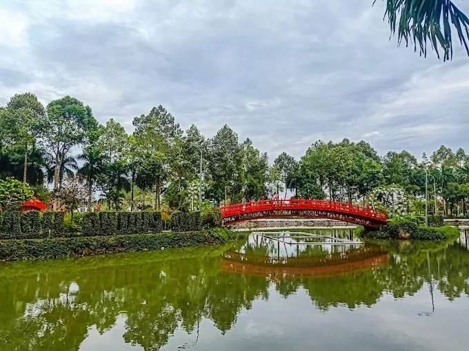 Cầu đỏ nổi bật trên hồ nước xanh, giữa khung cảnh cây xanh tươi tốt dưới bầu trời u ám.